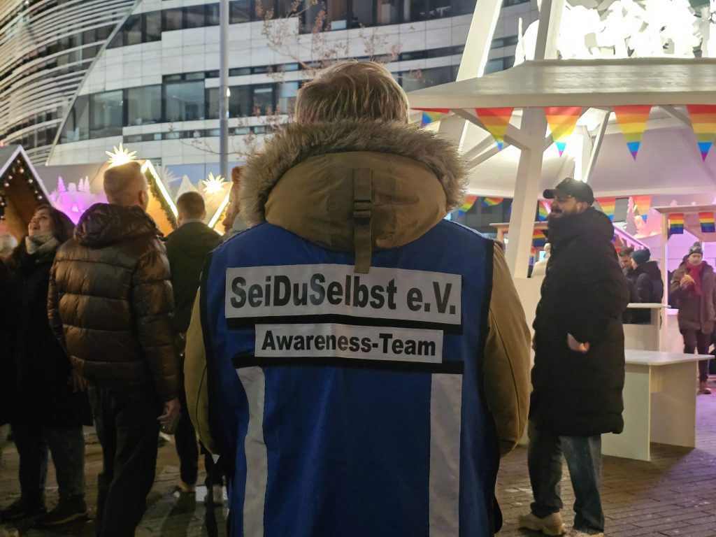 Mitglied des Awareness-Teams von SeiDuSelbst e.V. mit Rückenschild während einer Laufpräsenz durch die Besucherinnenmenge beim Pink Monday auf dem Düsseldorfer Weihnachtsmarkt 2025.